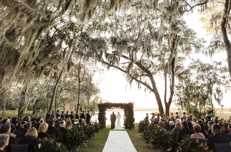 Outdoor wedding ceremony under branching trees with bride and groom facing alter | PartySlate