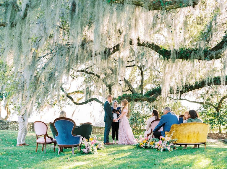 Intimate outdoor wedding ceremony shaded by branching tree with loved ones on plush sofas | PartySlate