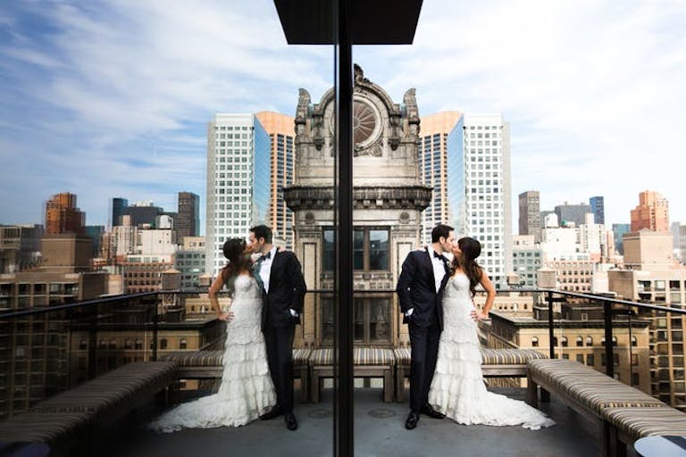 Bride and groom share a kiss atop NYC skyscraper with skyline in the background, mirrored by building | PartySlate