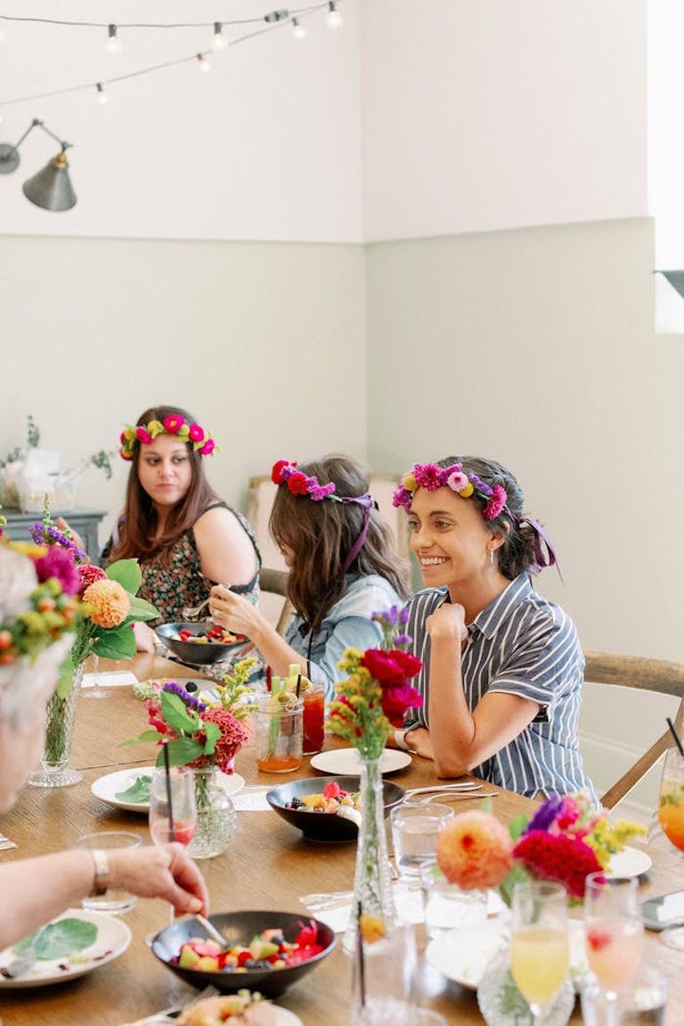 Baby shower event goers at table filled with colorful food don bright pink flower crowns | PartySlate