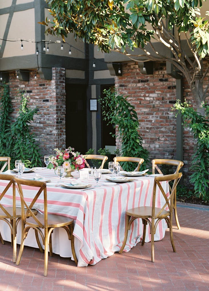 Banquet table with pink-and-white stripped tablecloth in courtyard at a wedding rehearsal dinner | PartySlate