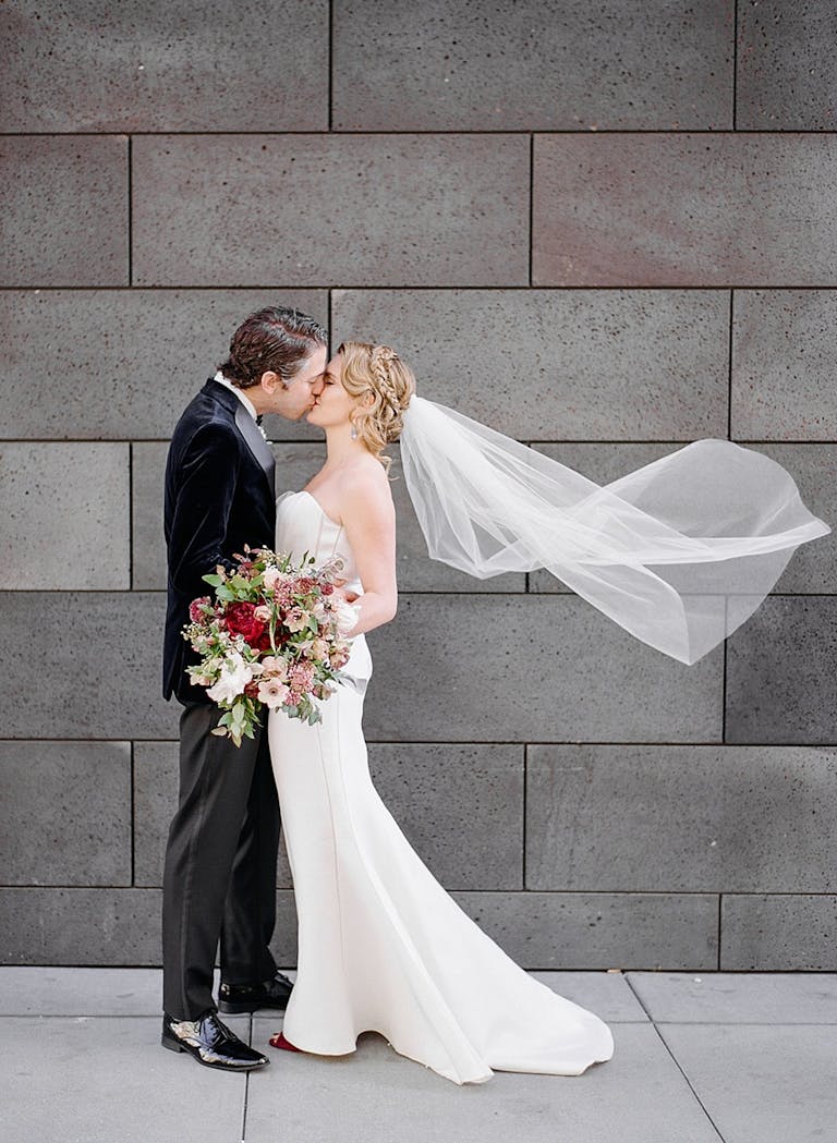bride with wind-blown veil and groom kiss in front of gray brick wall