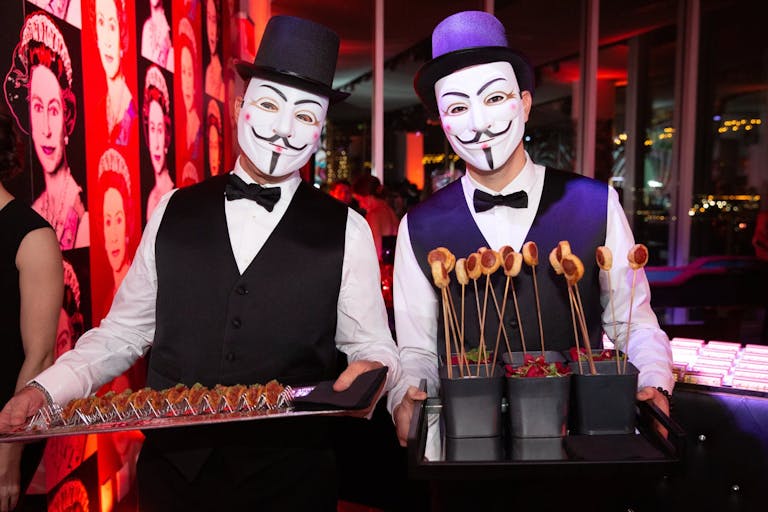 Two masked servers carrying trays of food