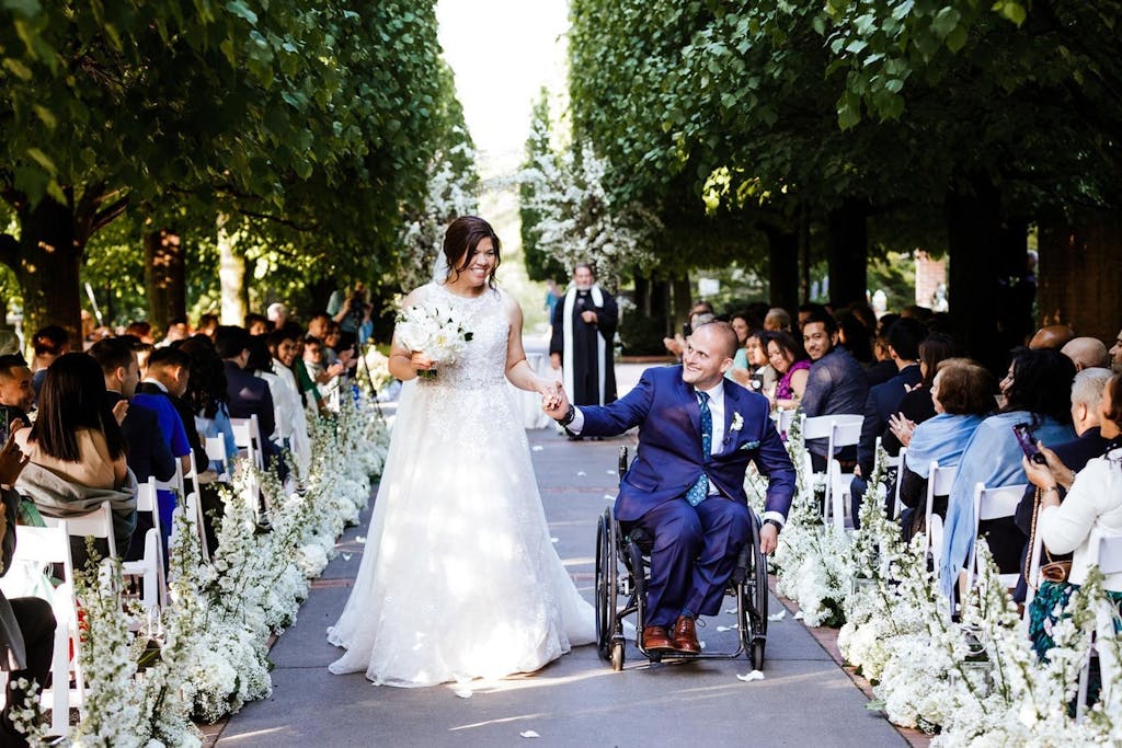 A bride and a groom in a wheel chair walk down the aisle. the aisle is lined with white florals and trees on the outside perimeter.
