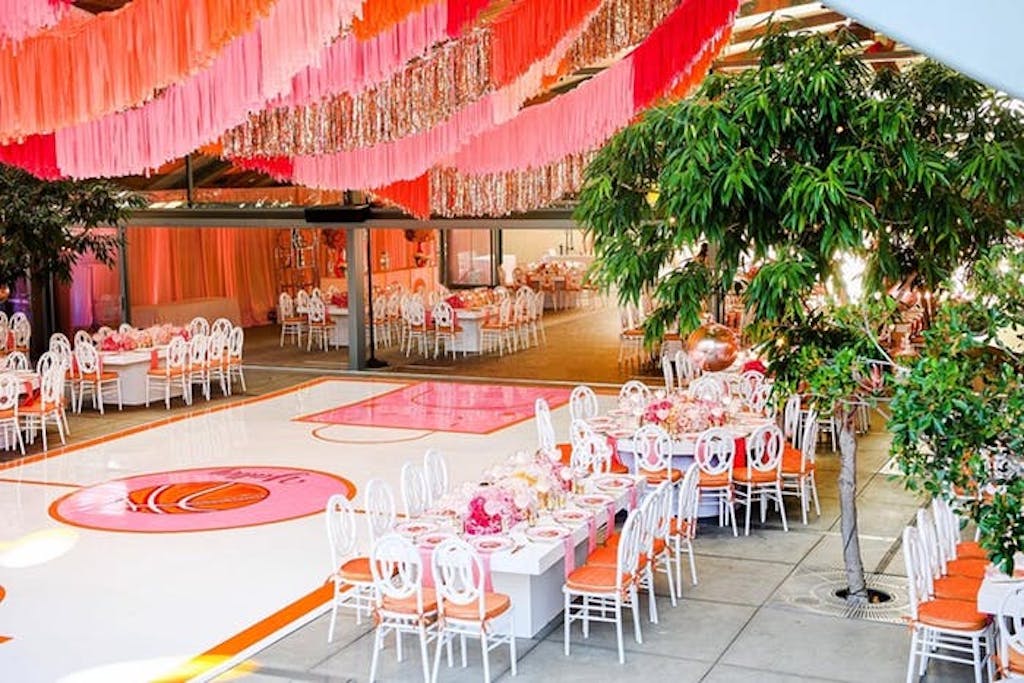 pink and silver tinsel hangs from ceiling above a white and pink basketball arena. Tables are placed next to the arena and green trees are around the perimeters
