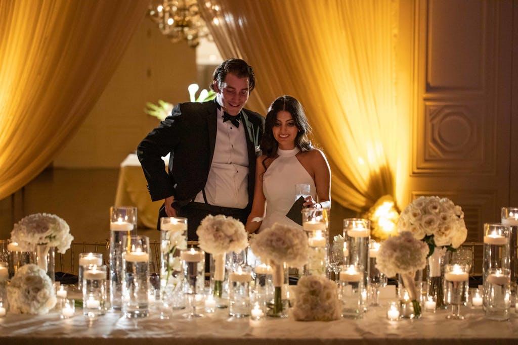 A bride and groom stand at the table admiring the centerpieces and decor. Dramatic up-lit draping is in the background.