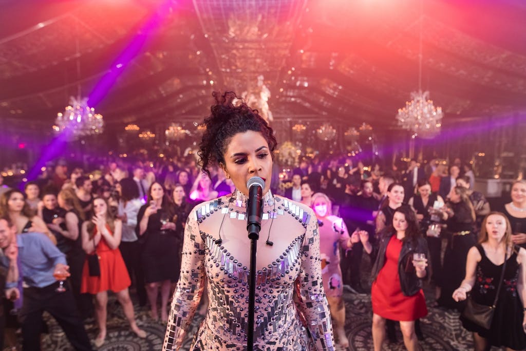 A singer stands on a stage singing with her back to the audience. chandeliers and pink lighting are in the background.