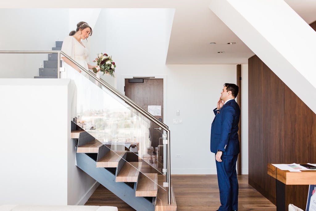 A bride walks down a minimalist stairway with the groom at the bottom wiping a tear.