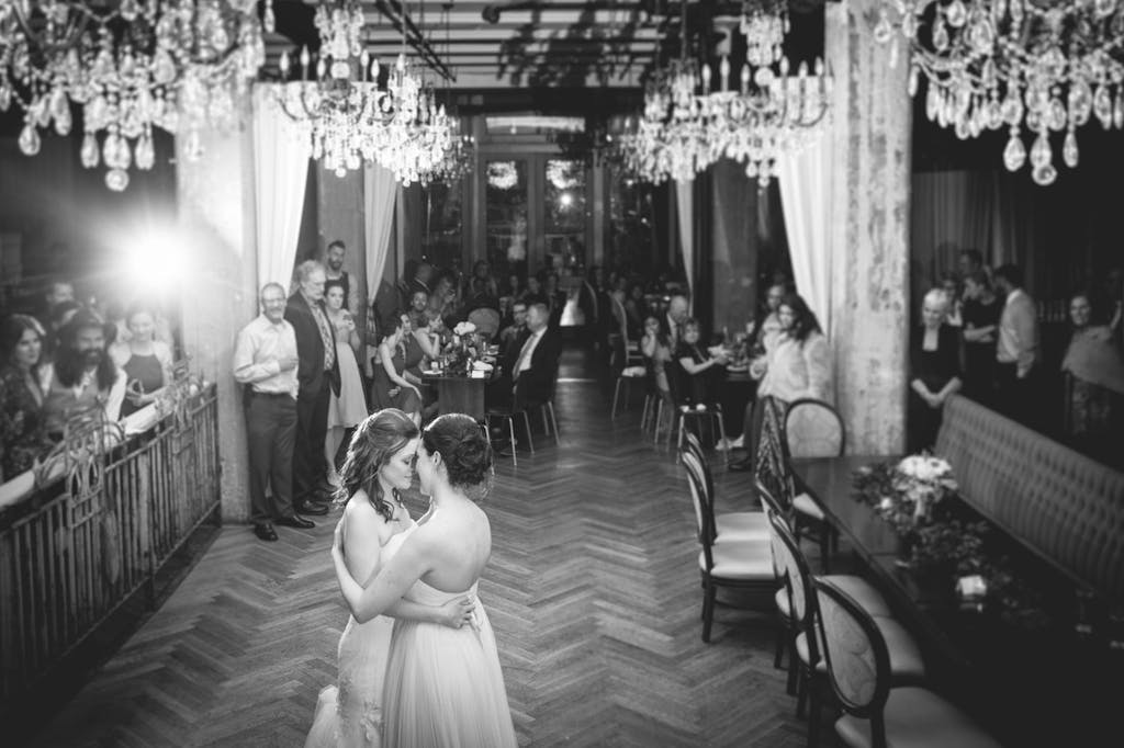 Two brides are on the dance floor with chandeliers above. Wedding guests surround the area and watch the couple.