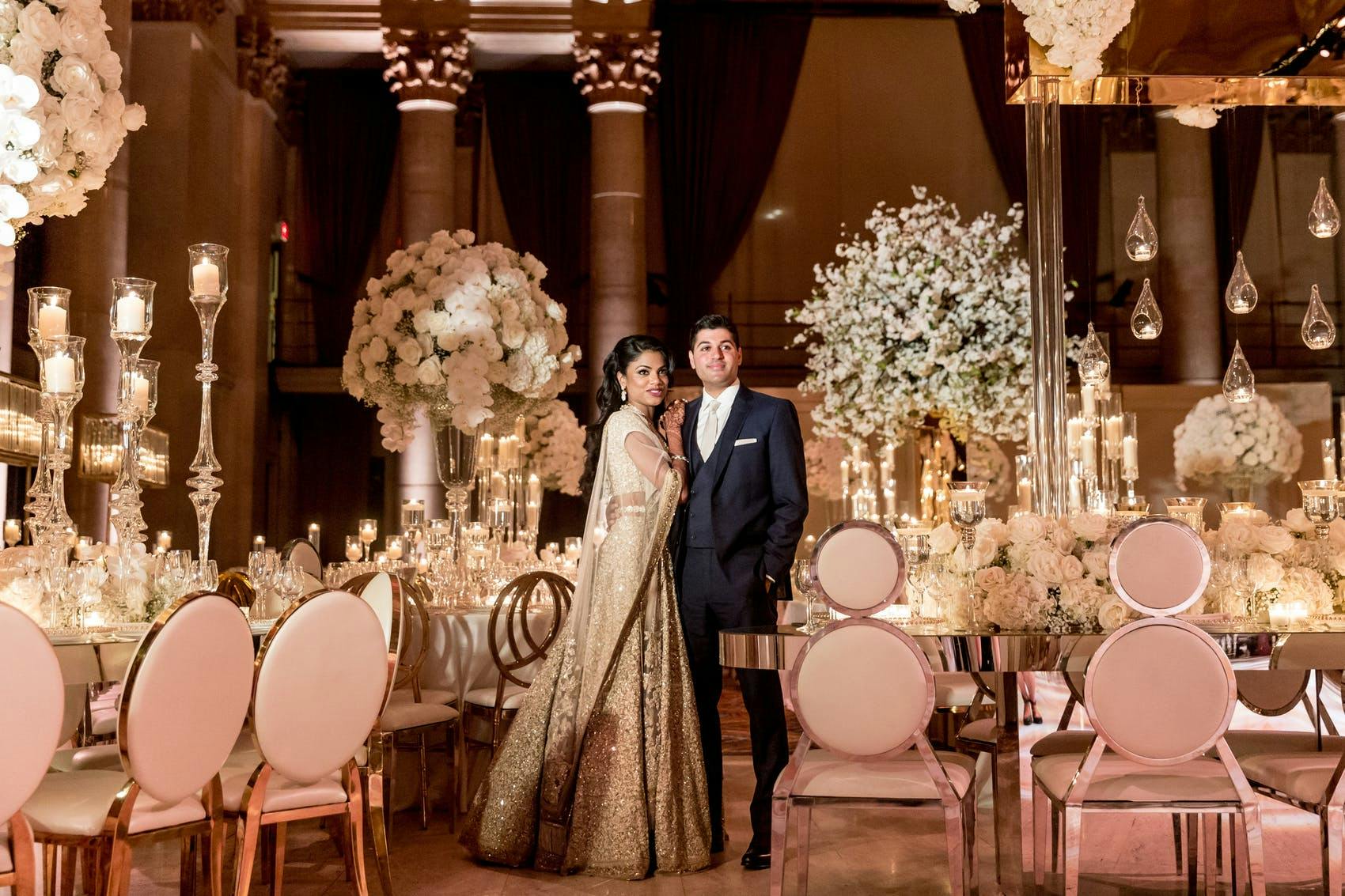 A husband and wife stand between white tables with pink chairs and large white bouquets.