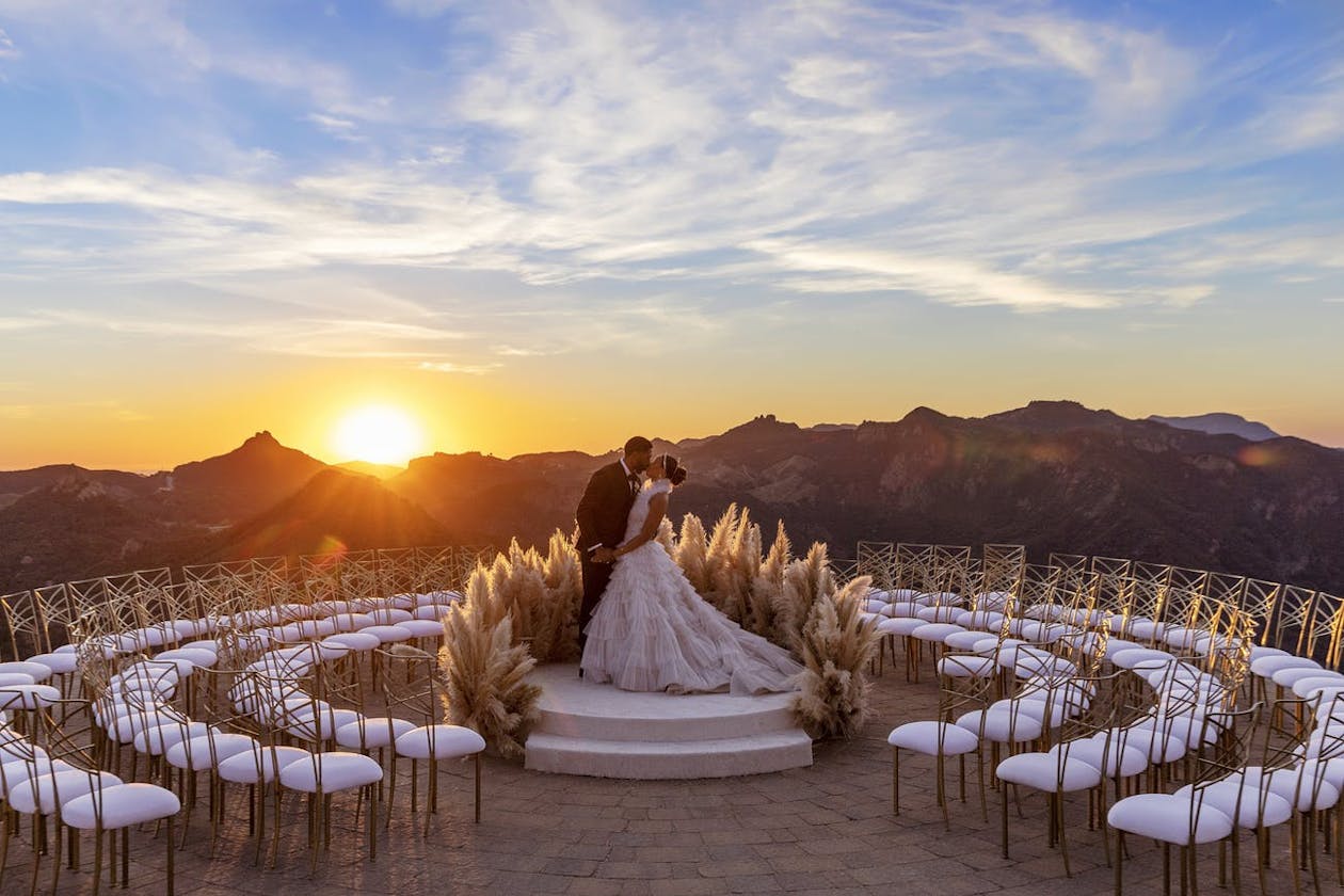 bride and groom kissing against backdrop of sunset, mountains, and ceremonial seating