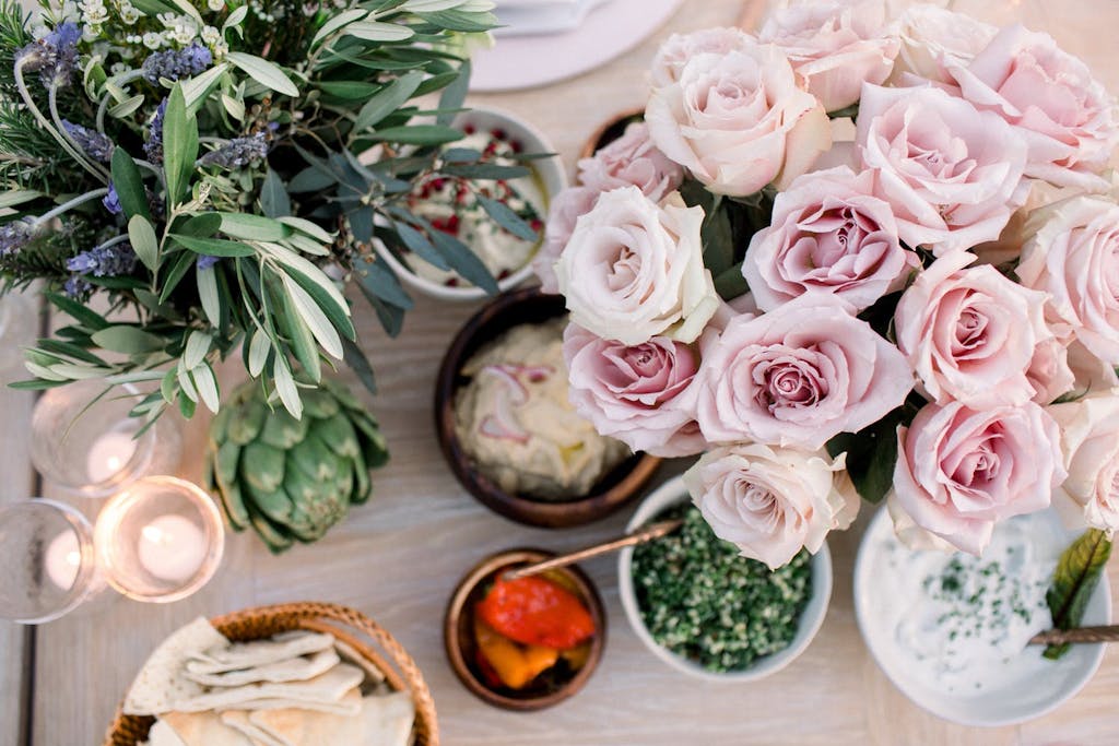 still life photograph of blush-colored roses, sage, artichoke, votive candles, and bowls of pita and various spreads
