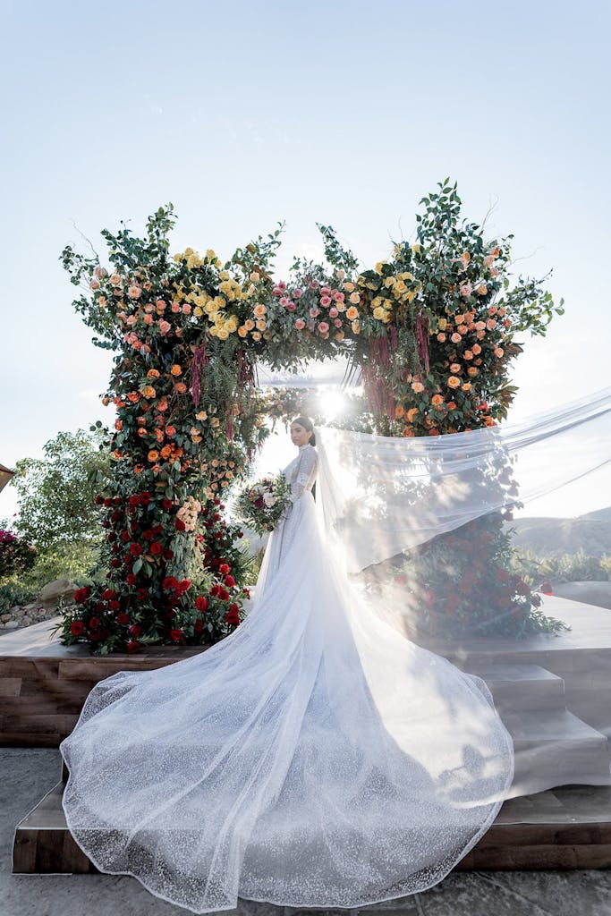 bride with windswept veil stands in front of a bloom-covered ceremonial arch and setting sun