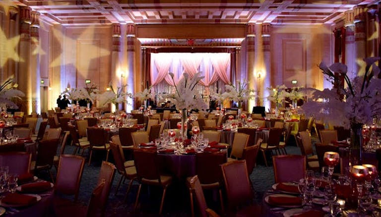 Large room with red tint and chairs and tables all around with curtains and stage at the end