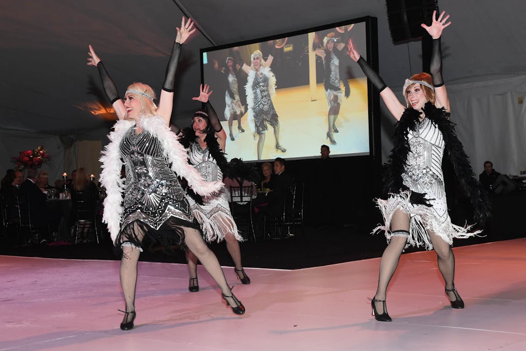 Three flapper dancers strike a pose in front of giant television showing their live performance.