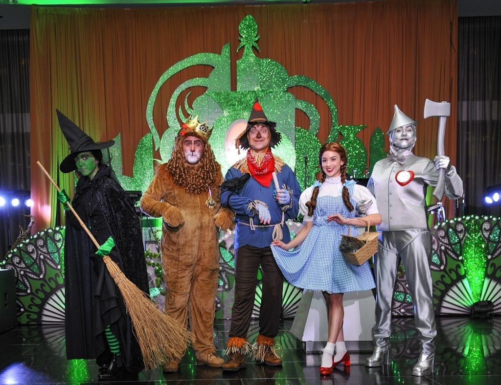 People pose as Wicked Witch of the West, the Cowardly Lion, the Scarecrow, Dorothy, and the Tinman in front of an Emerald City cutout.
