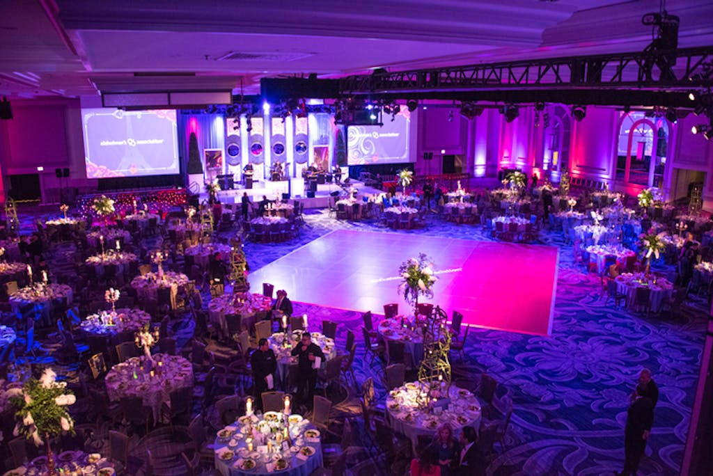 Industrial ballroom with banquet tables, dance floor, and pink/purple uplighting.