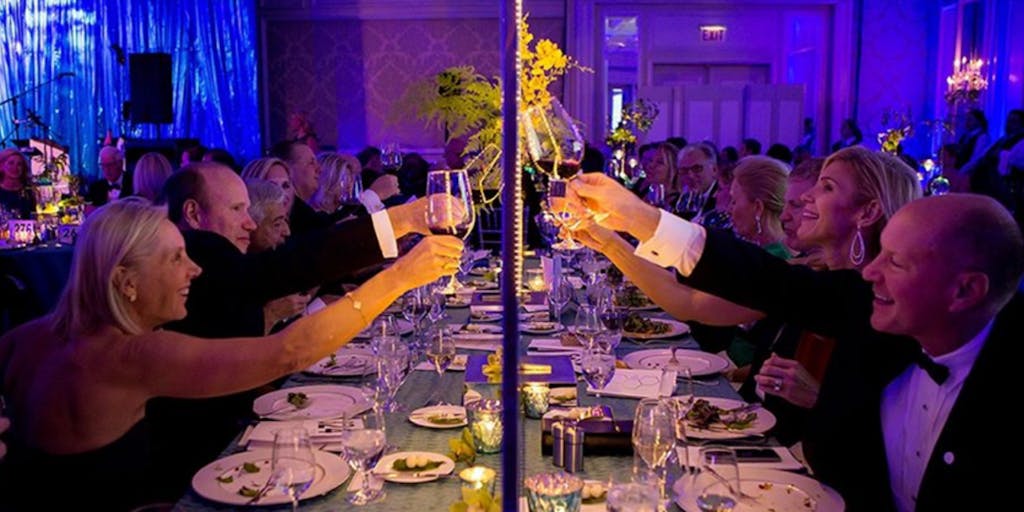 Guest cheers toward center of rectangular banquet table against backdrop of royal blue uplighting