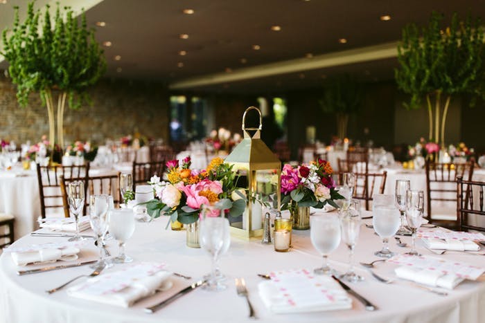 A round table with white linen and place settings. Multicolored florals surround a golden lantern with a candle inside.