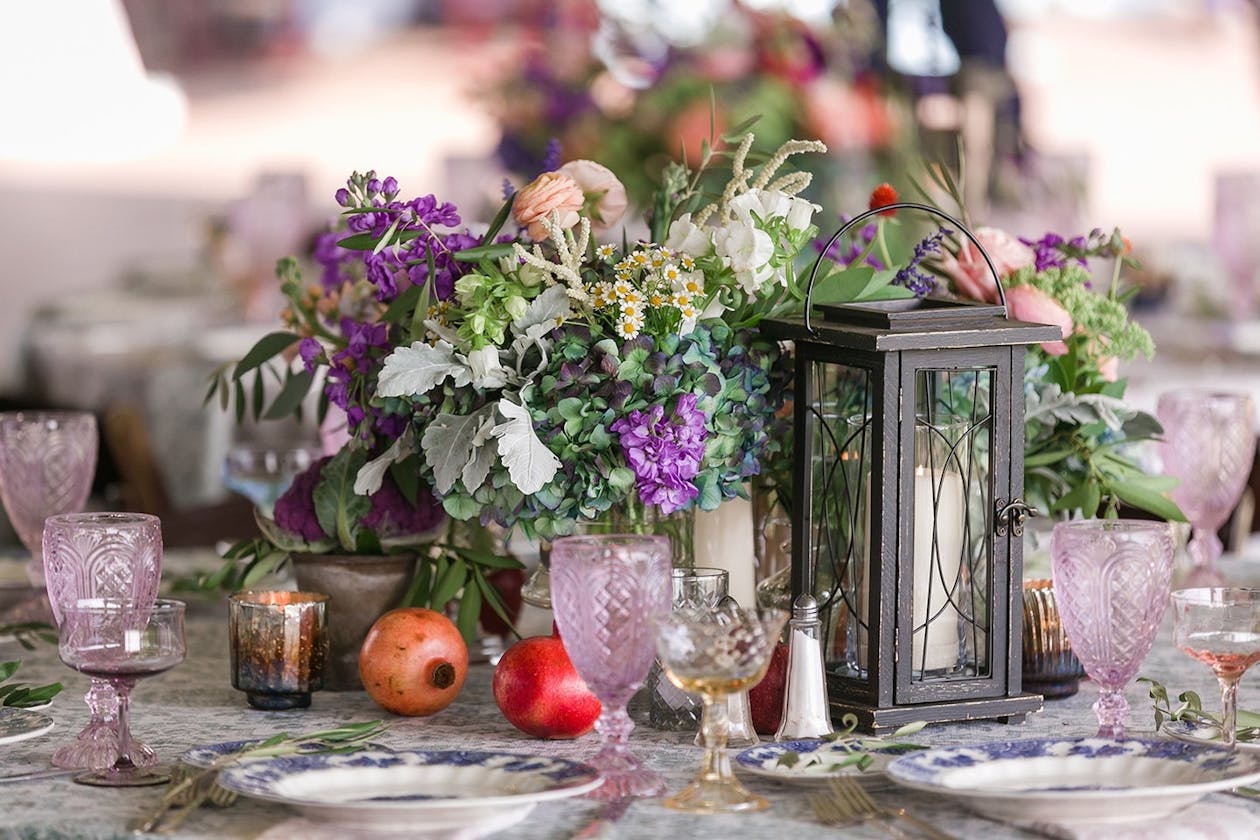A centerpiece with purple and red florals an sprawling green plants next too candles in lanterns