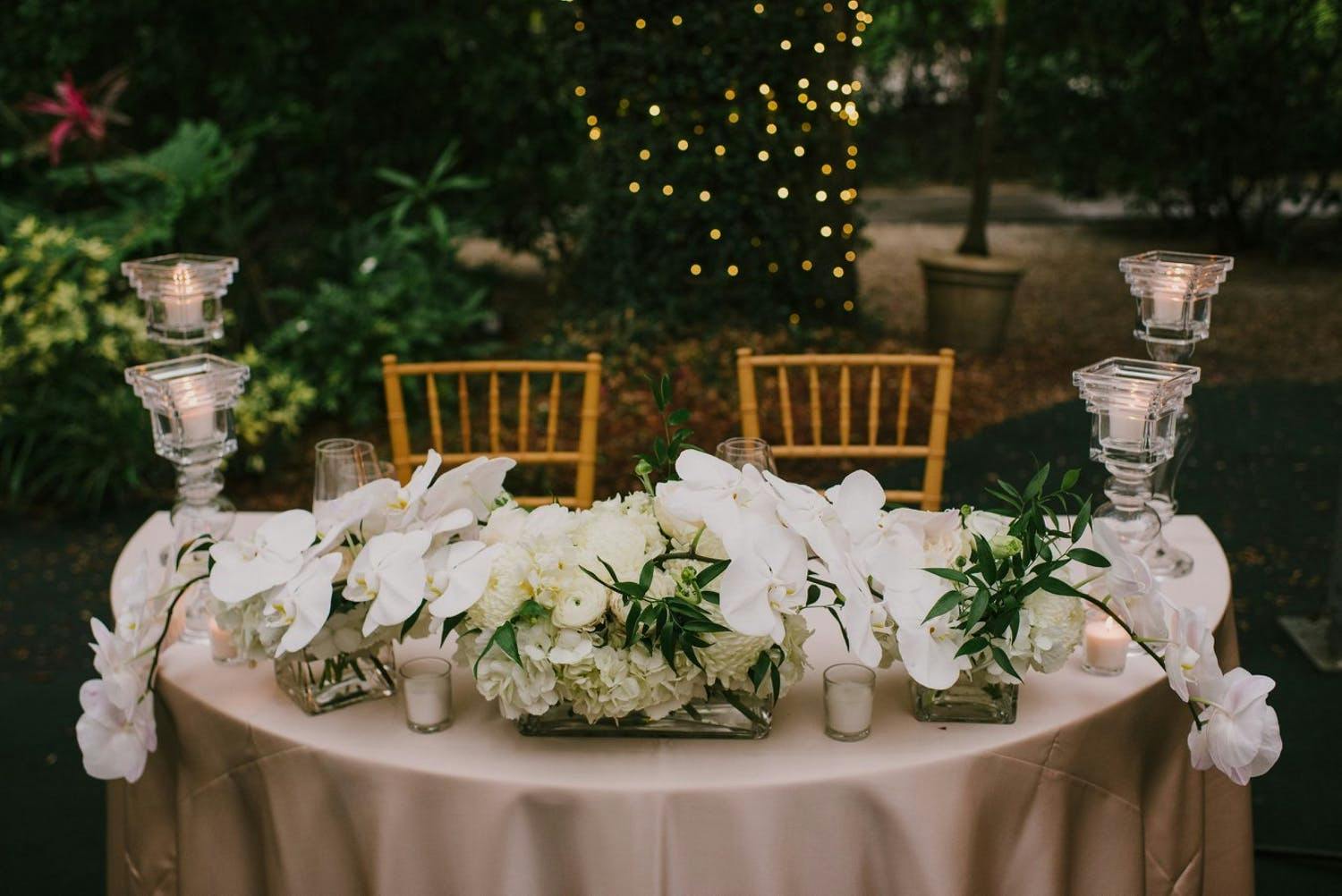 Sweetheart table with pale pink linen, cascading white orchid wedding centerpiece and four glass candle pillars. In the background is greenery and a tree trunk wrapped in string lighting.
