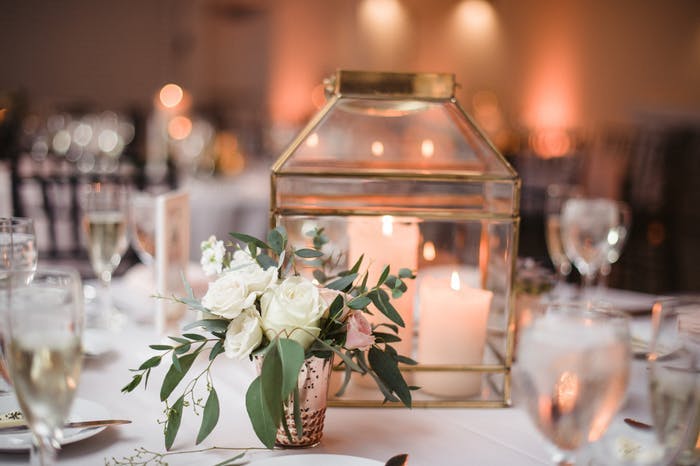 multiple white pillar candles are in the middle of a wide transparent lantern. White florals and greenery are in a votive sized vase interspersed with the lanterns