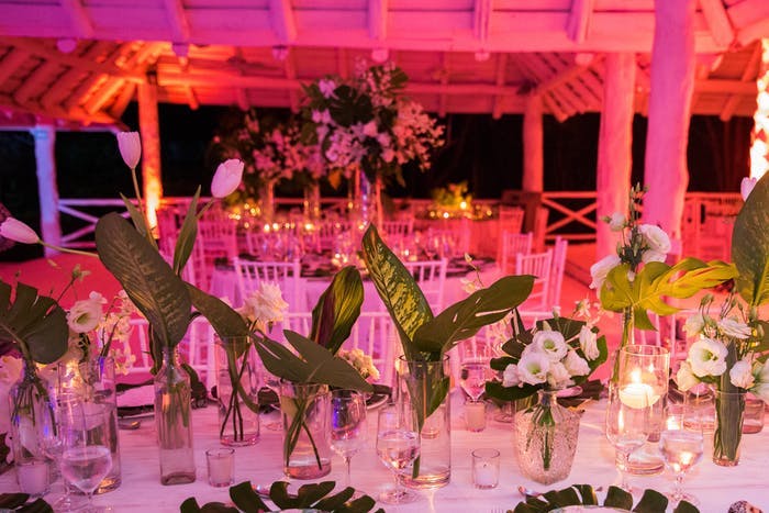 A large gazebo washed in pink light with round tables. Centerpieces are short glasses with leafy greens and small florals