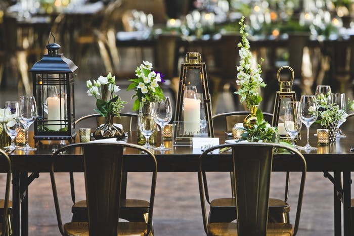 A clean metalic table and chair combination with varrying heights and styles of lantern. Mixed into the lanterns are varying styles and heights of white and green floral arrangements.