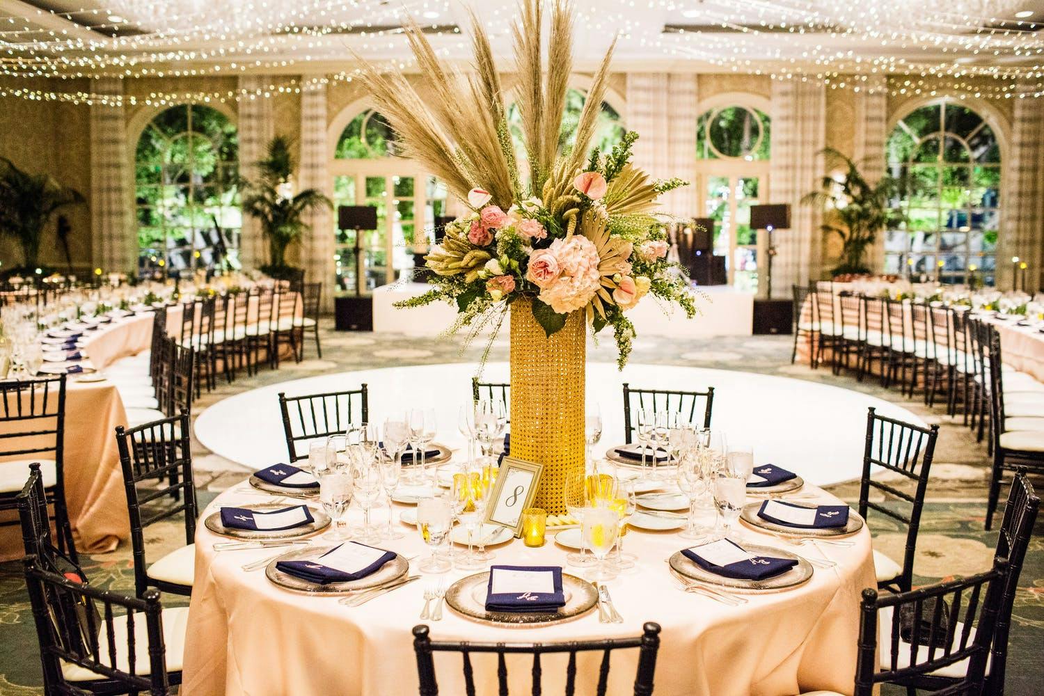 Ballroom with exposed brick and floor to ceiling windows in backdrop. Forefront of photo shows a round banquet table in peach linen with a boho-vibe centerpiece consisting of a wicker vase and pampas grass with blooms.