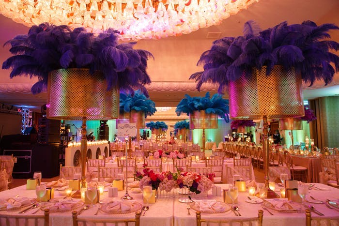 A large room with a light wash of pink and centerpieces in the center of rectangular tables that resemble table lamps with purple feathers coming from the top