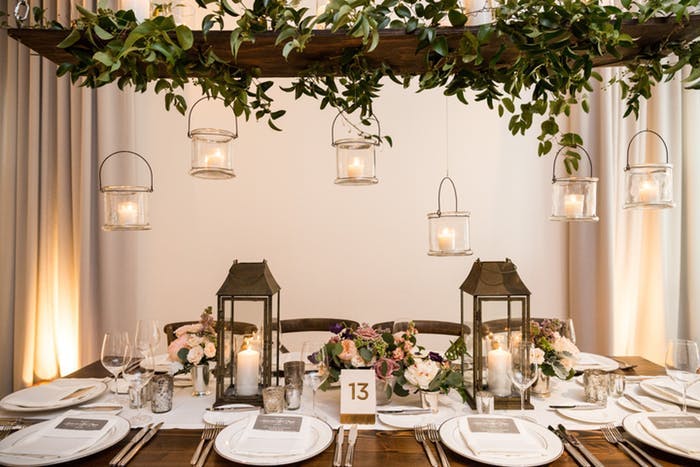 A rectangular table with place settings and low floral arrangements down the center with two lanterns on either side. A rail covered in green leaves is above the table and candles in transparent buckets hang at varying heights