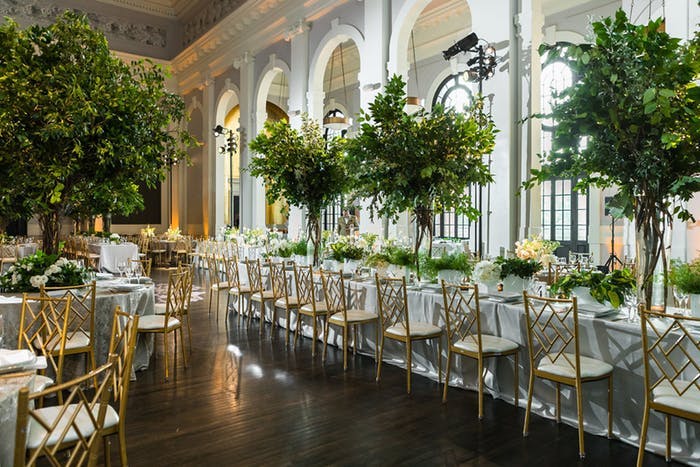 light-filled arched windows beside long rectangular tables with white linens. Trees are planted in the middle scattered through the tables and dark floors