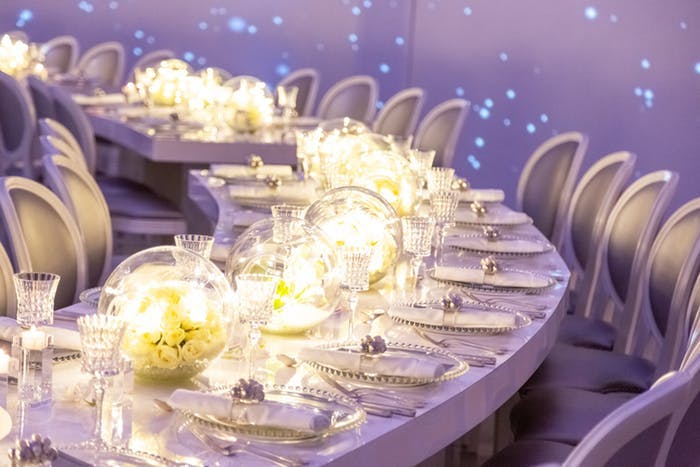 Walls with blue laser beams behind a white circular tables. Transparent globes are filled with gold and yellow lighting and white florals