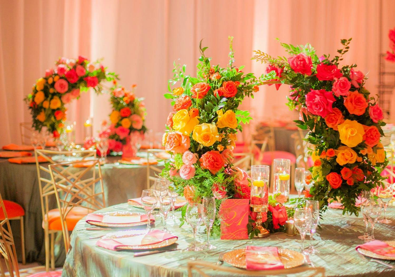 Two banquet tables with pale green linens and pink, yellow, red flower-studded wreath centerpieces against a backdrop of pale pink drapery.