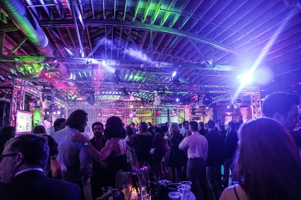 Industrial gala space filled with guests and green and purple uplighting.