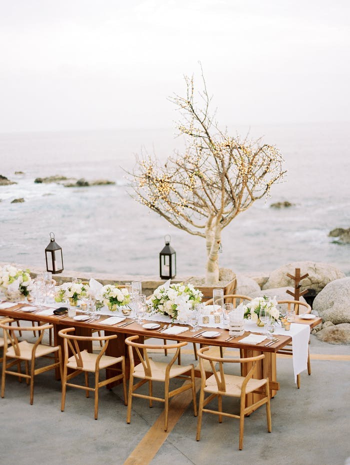 A light wooden rectangular table overlooking choppy water with white linen table runner and low green and white florals. A branchy tree sans leaves sits between the table and the water and metal lanterns hang above the table on a string.