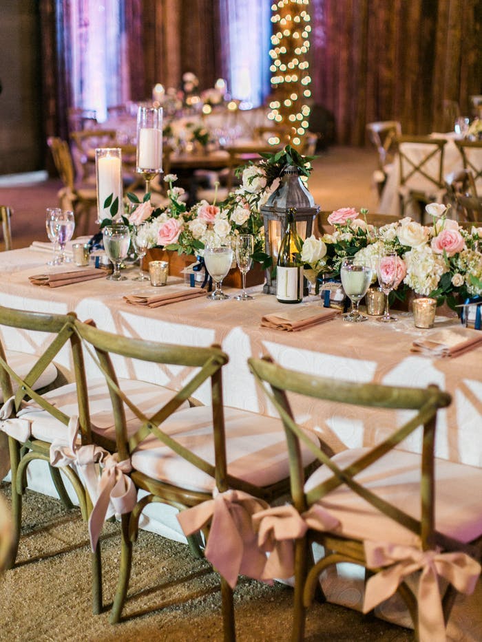 Rectangular table with white linen and wooden chairs surrounding. A lantern is in the middle of the table decor surrounded by pink and white flowers and greenery.