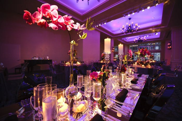A dimly lit room with purple uplighting, glowing candles, and pink orchid wedding centerpieces.
