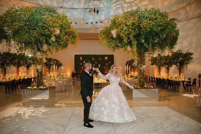 a couple walks through their reception room hand in hand. The room is filled with green trees and warm lighting
