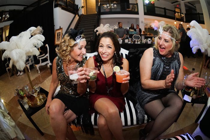 Three Women Sit on a Couch with 20s Themed Outfits and Flanked by Feathery Décor | PartySlate