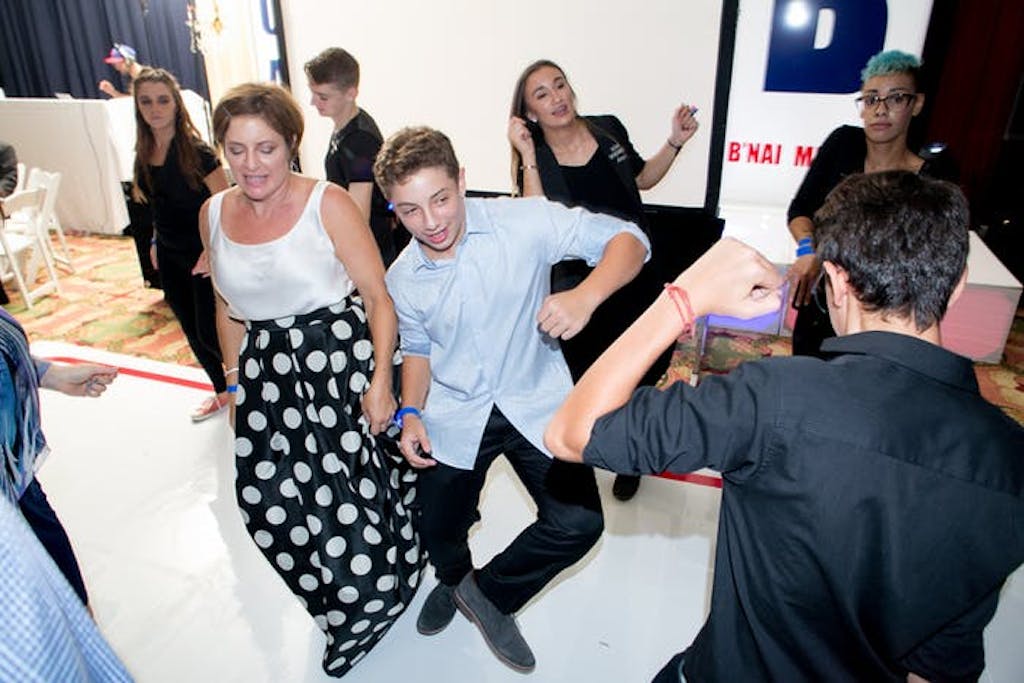 A mom and son dance together on an all white dance floor. Party guests are dancing around them