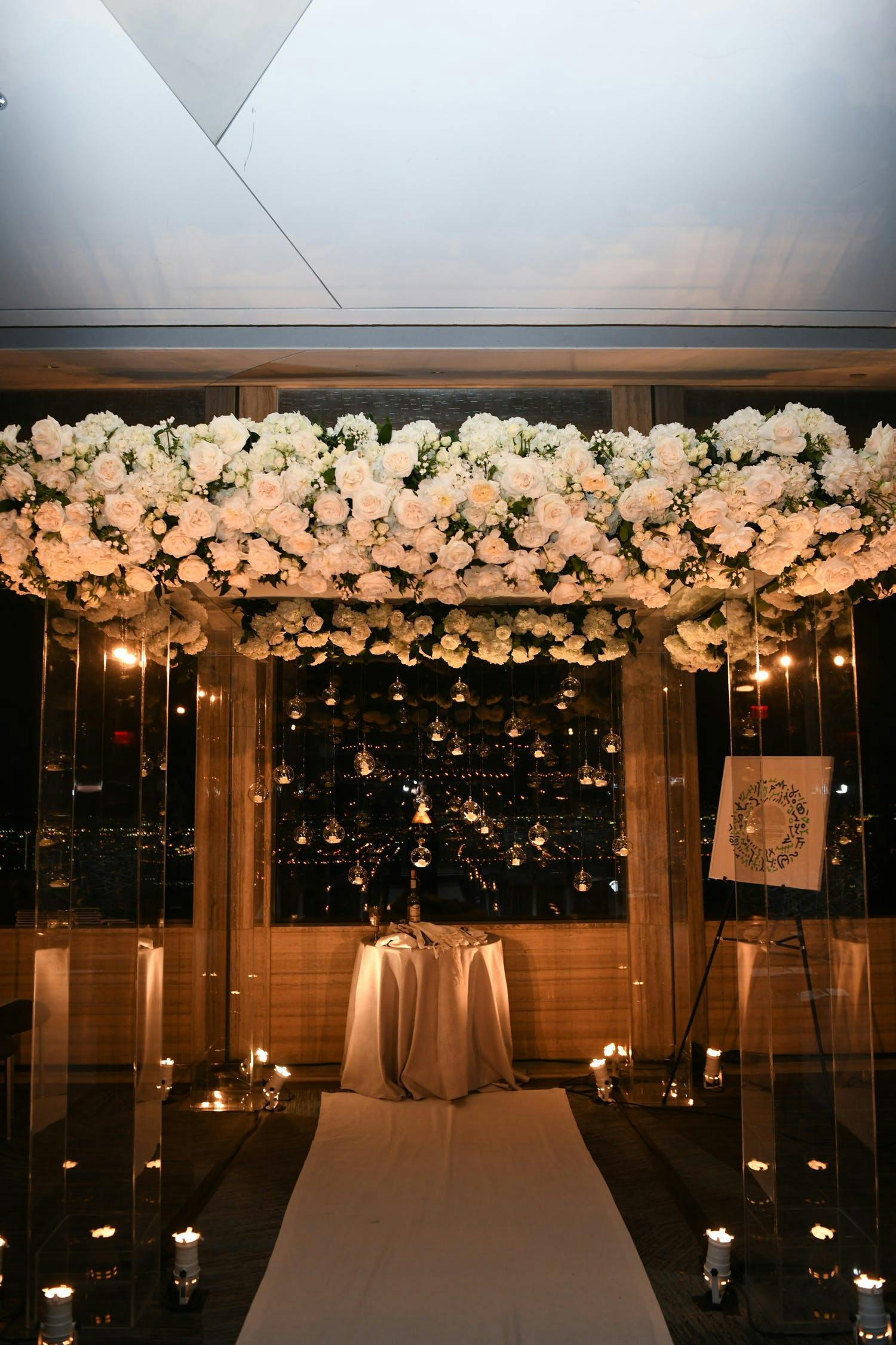 Candlelit ceremony altar with night city view in backdrop. The altar's roof is covered in clusters of white blooms intermixed with greenery.