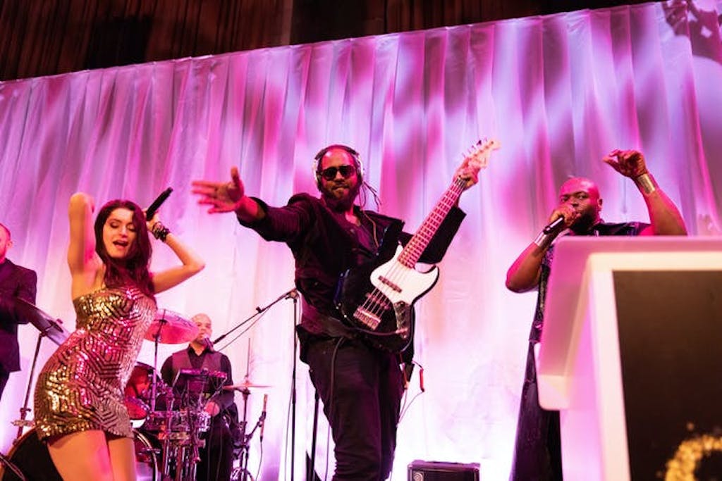 A man stands with one arm reached out to the audience and the other on an electric guitar. A lady in a gold tight dress dances to the left. A pink wavy backdrop is behind them