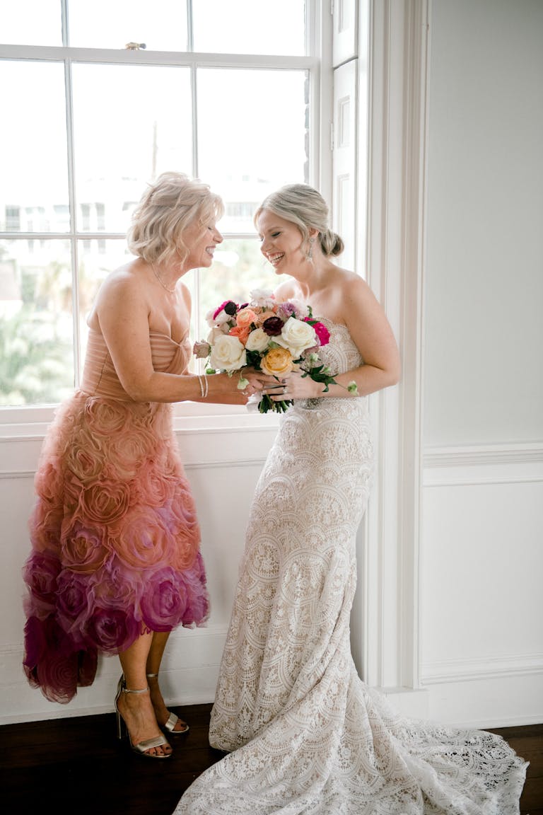 A bride and her mother share a laugh while both holding her white, peas, and pink, rose bouquet.