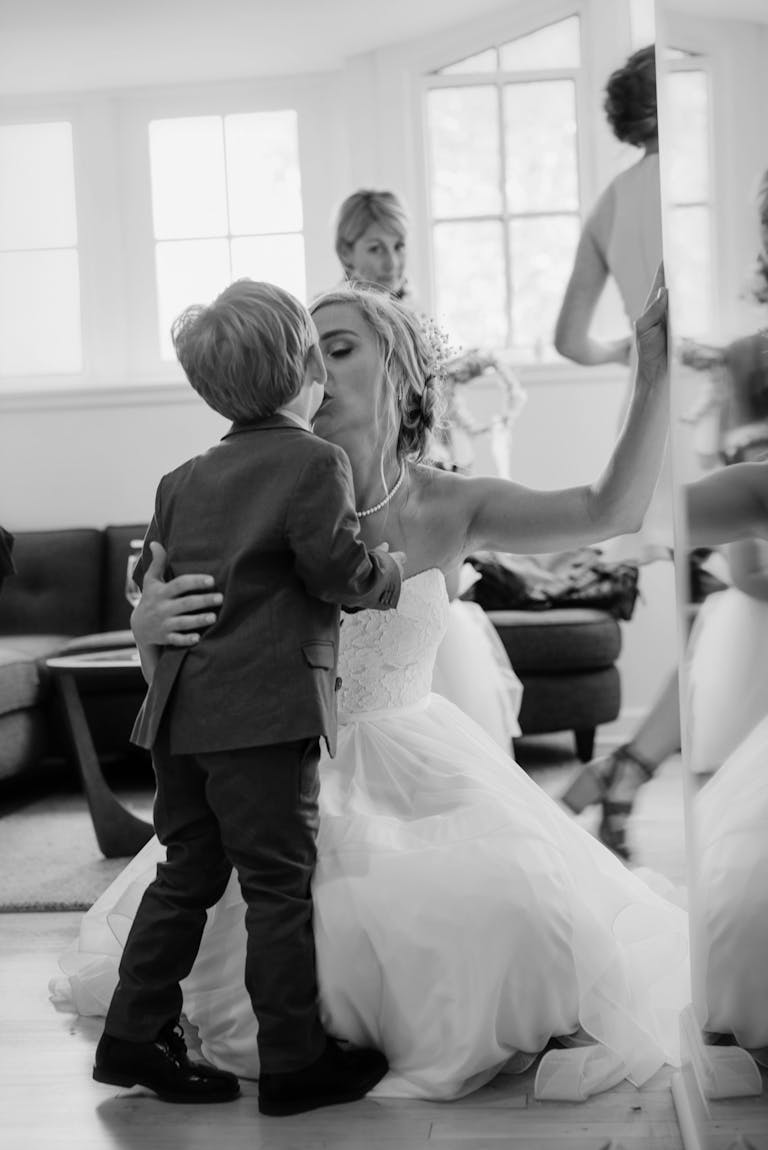Black and white photo of bride in white wedding dress kissing young son who is wearing a tux.