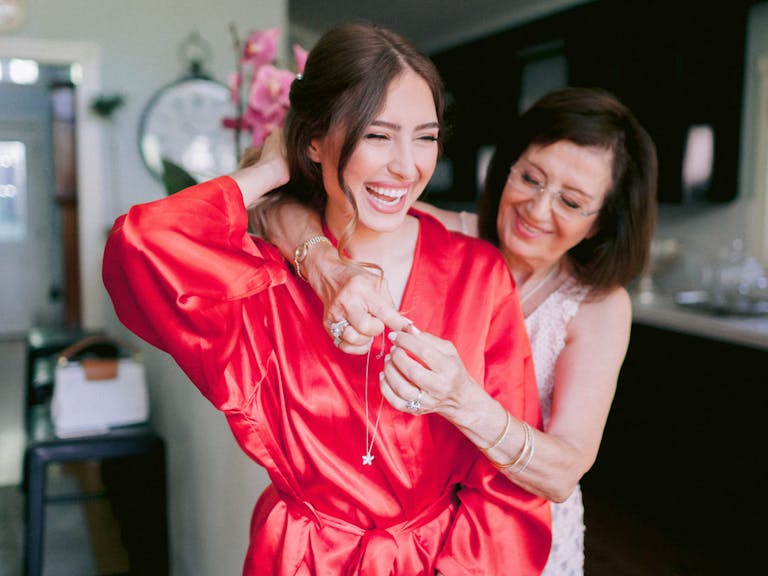 A bride in a coral-colored robe laughs as her mother stands behind her and helps clasp her necklace.
