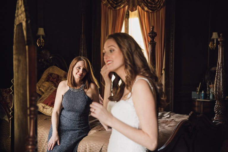 Mom sits on bed and watches daughter put on earrings before wedding ceremony.