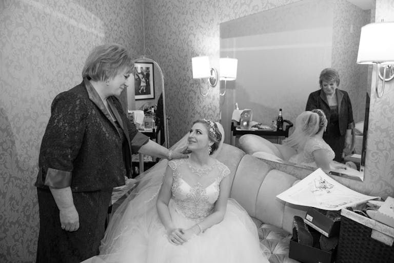 A bride sits in a dressing room and looks lovingly up at her mother who touches her on the shoulder.