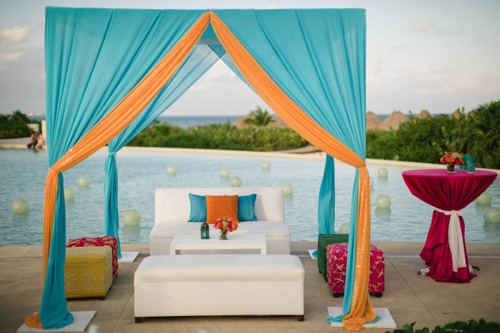 White couch under a blue and orange canopy in front of the water. Pink covered cocktail tables are off to the sides