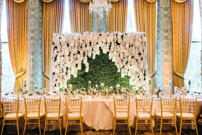 windows with gold drapes behind a rectangular table. A greenery backdrop is behind the table and is flanked with white flowers